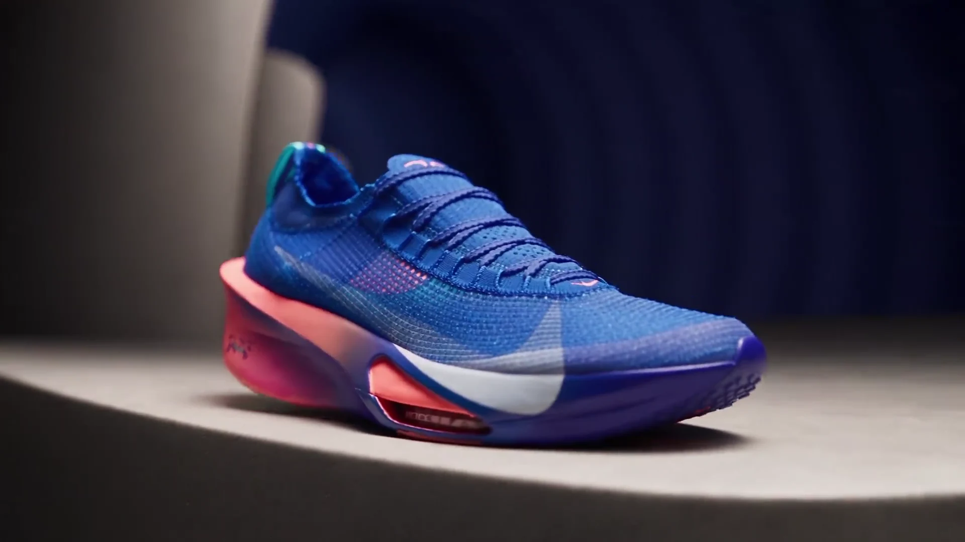 A blue knitted running shoe with a pink sole on a studio platform